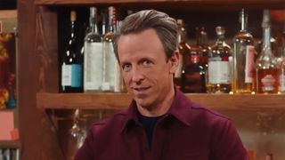 Seth Meyers stands behind a bar and stares straight at the camera. He is wearing a maroon button down shirt.