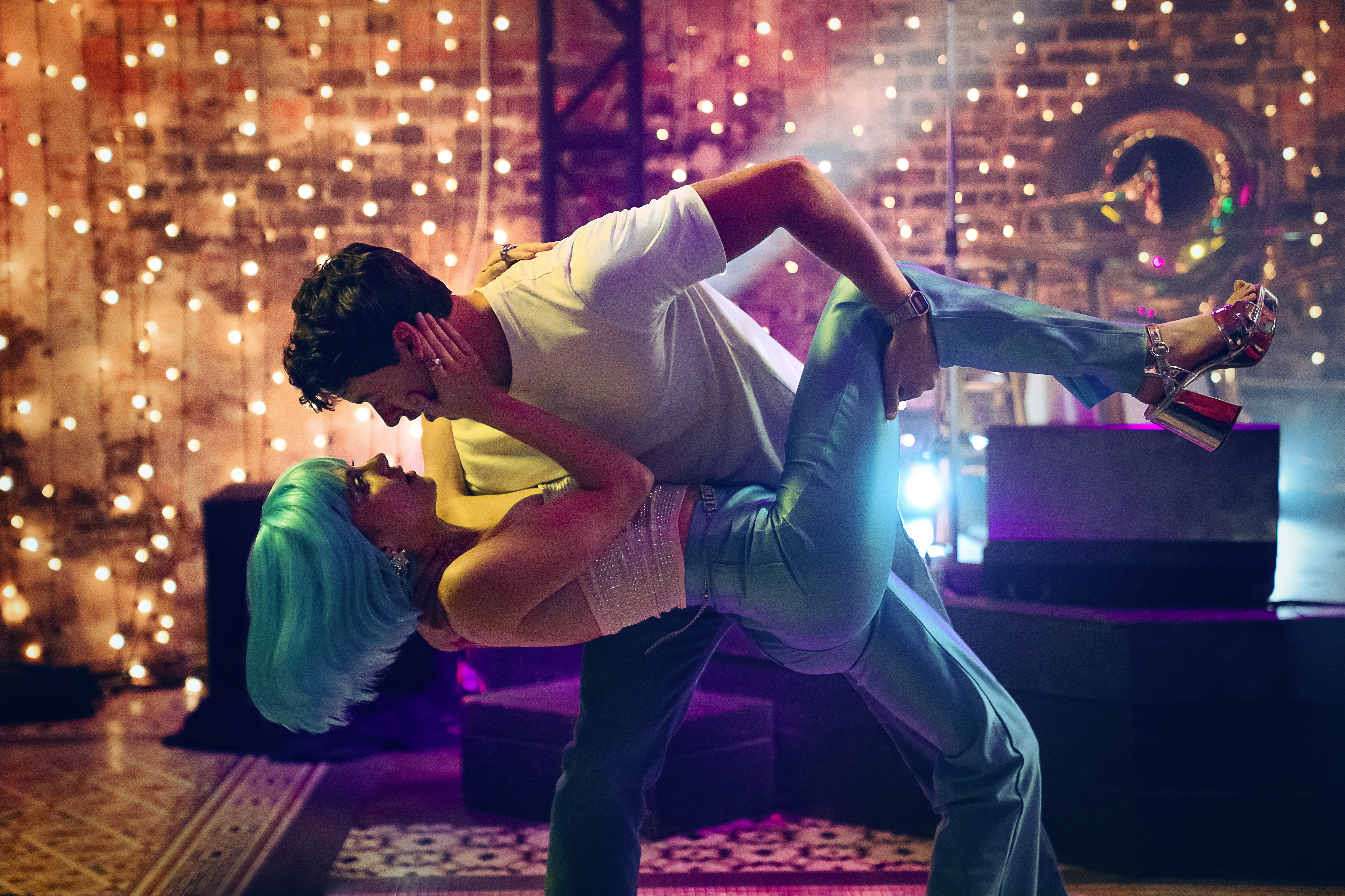 A man in a white T-shirt (Tom Blyth as Alex) dips a woman wearing a blue wig (Emily Bader as Poppy) on a dance floor. Behind them, a stage and brick wall illuminated with strands of twinkle lights, in the Netflix movie 'People We Meet on Vacation.'