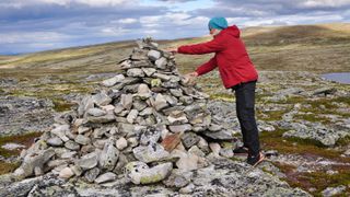 A hiker in a red jacket places rock on a rock cairn on a moor