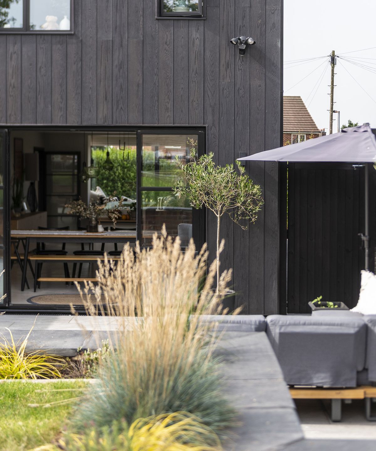 The rear of a contemporary renovation project taken from the garden. Tufts of ornamental grass can be seen as well as a patio leading out from the open glass patio doors