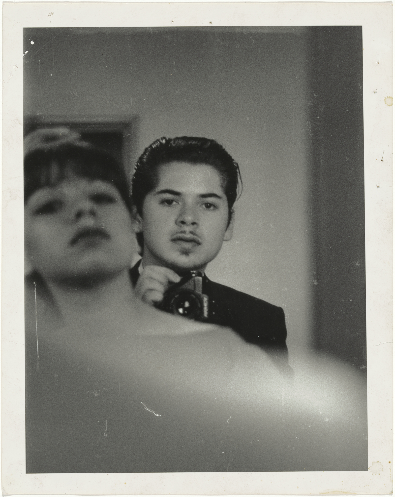 Black and white self-portrait reflected in a mirror, showing a young man holding a camera beside the blurred face of another figure.