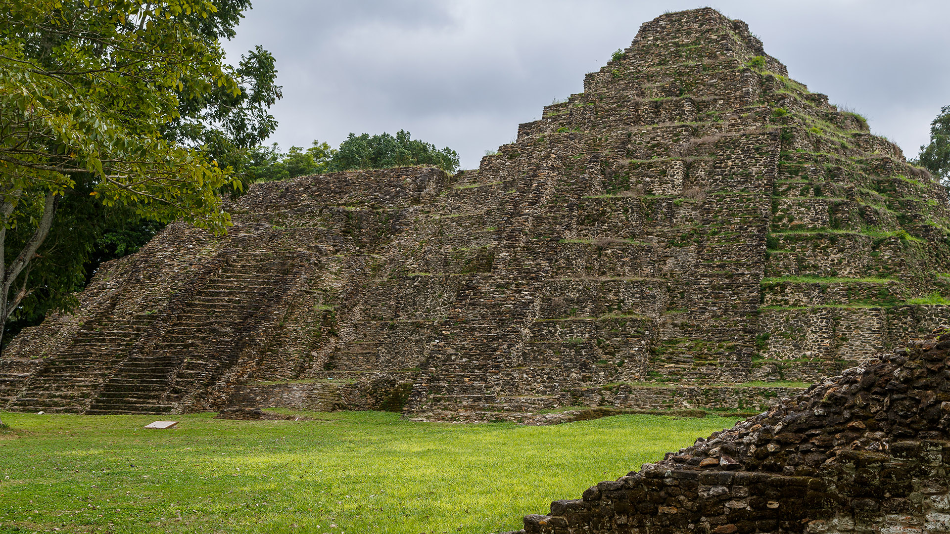 We see the ruins of a stone pyramid on a grassy field near some trees.