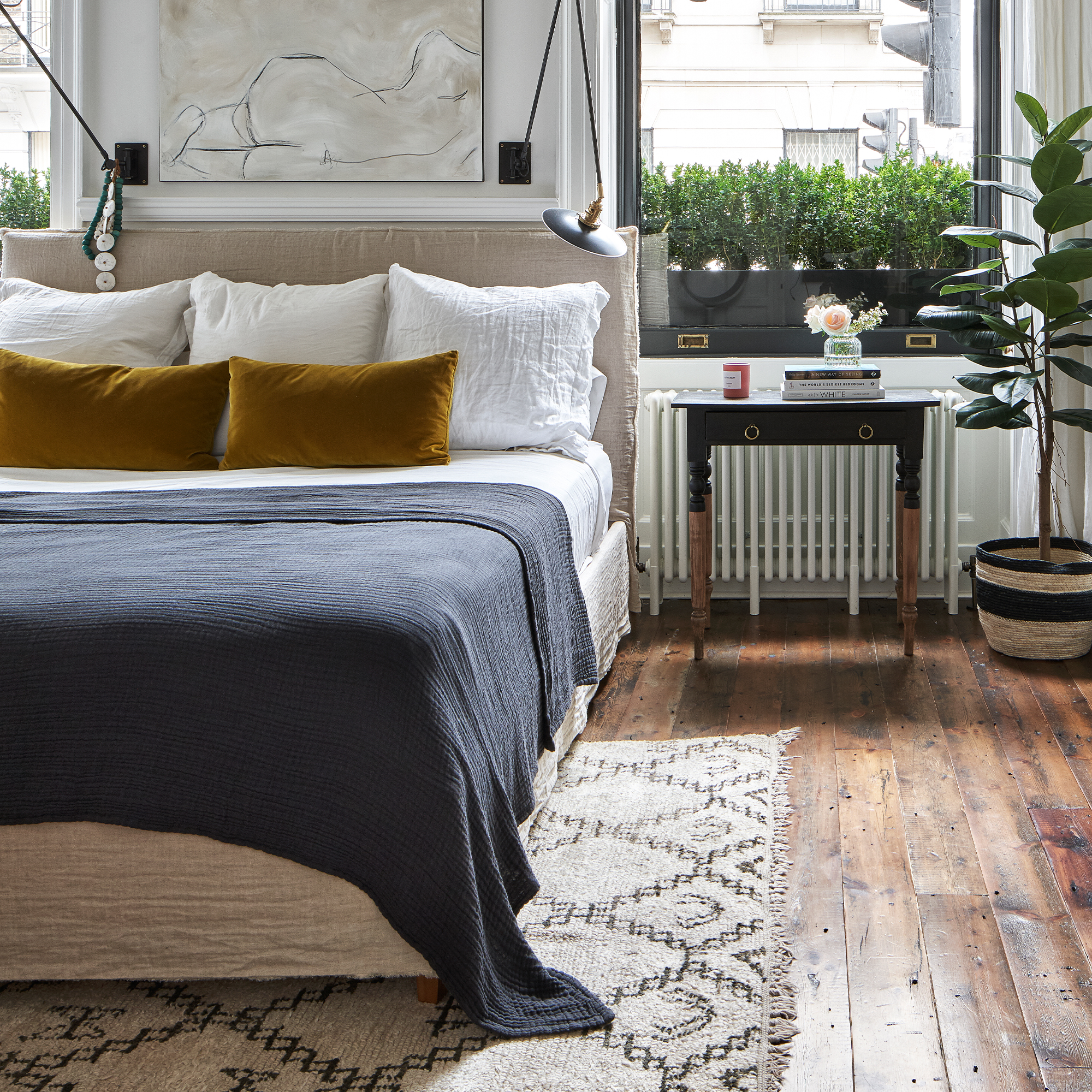 A bedroom with two large windows on the wall behind the double bed which is dressed in white bed linen with mustard yellow velvet cushions and a navy throw