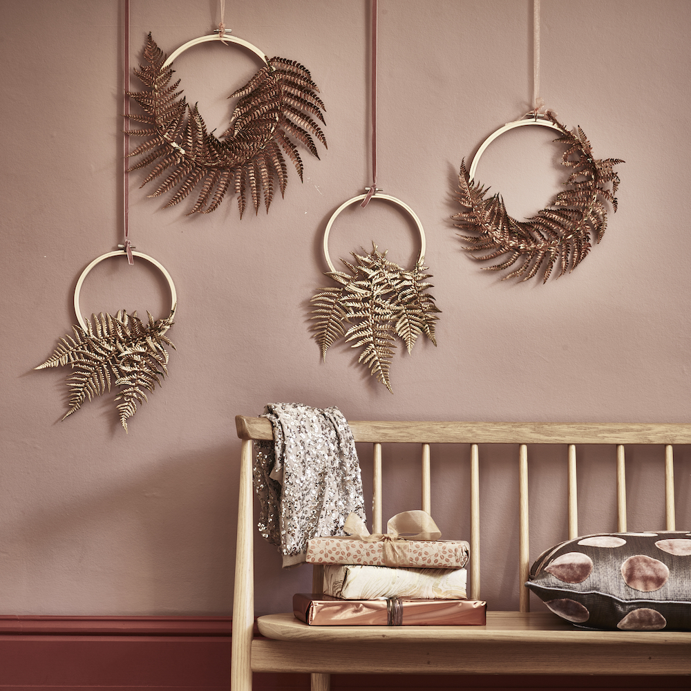 Gold rings adorned with fern leaves and sprayed in metallic spray, hung against a pale pink wall above a simple wooden bench