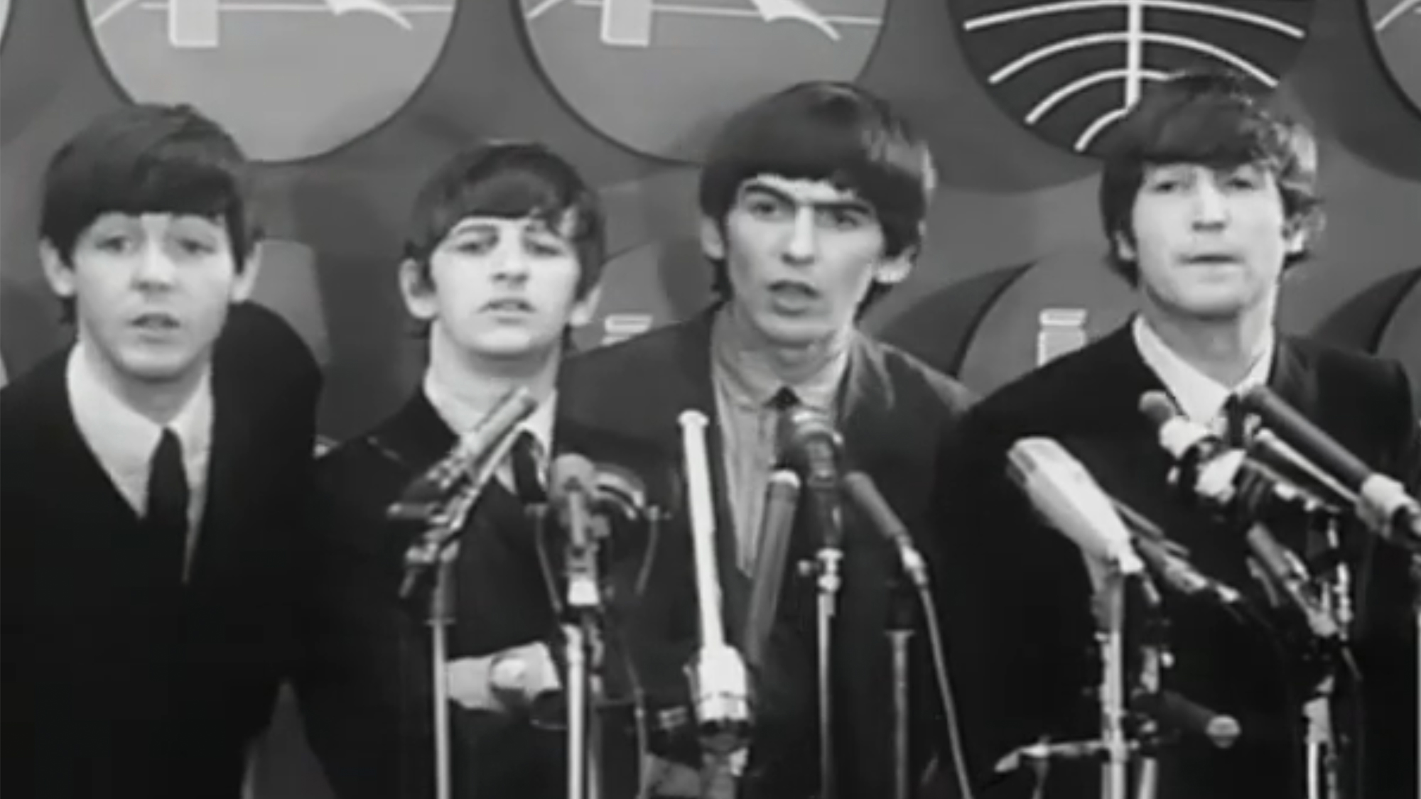 The Beatles speaking into microphones at their press conference at an airport in 1964