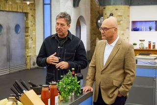 John Torode and Gregg Wallace standing at the edge of a workstation in the Celebrity MasterChef kitchen, talking to an unseen contestant