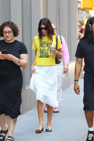 Kaia Gerber wearing a yellow graphic T-shirt with a white midi skirt and ballet flats in NYC.