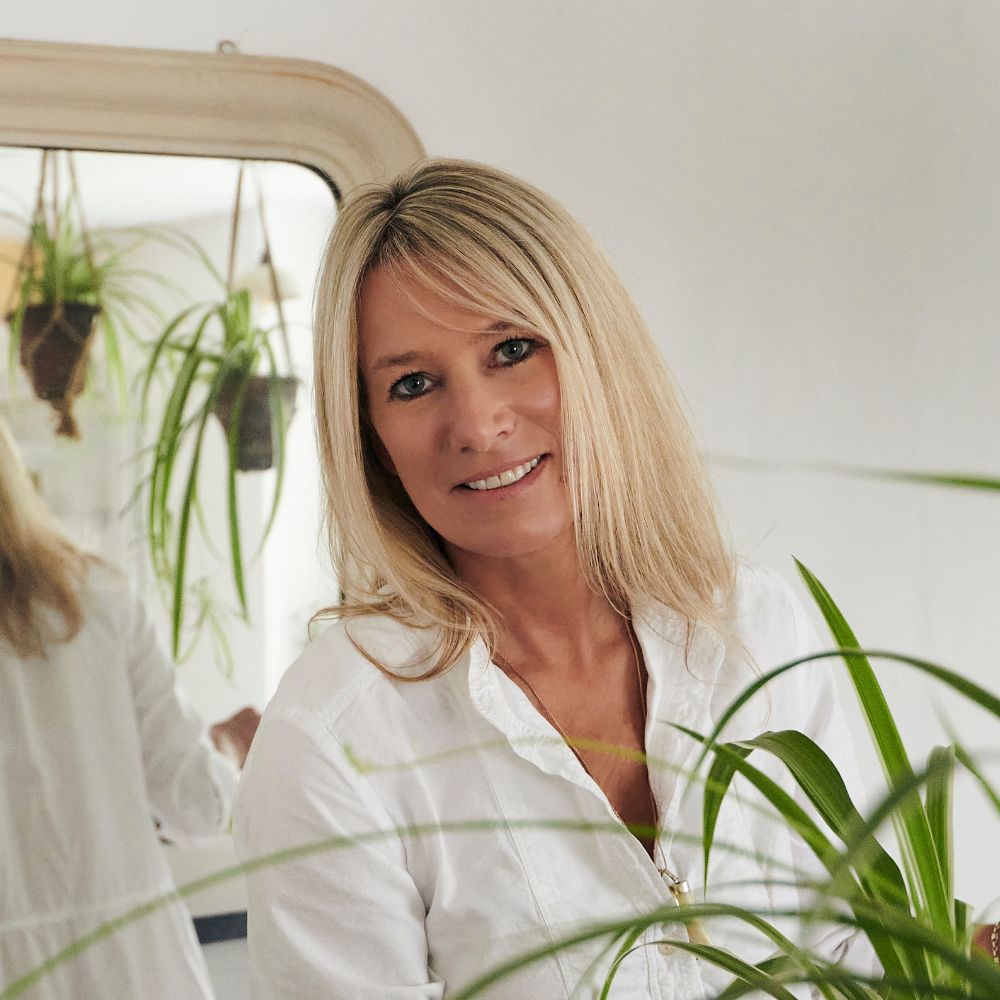 A blonde woman in a white shirt, with houseplants in the foreground and a mirror in the background 