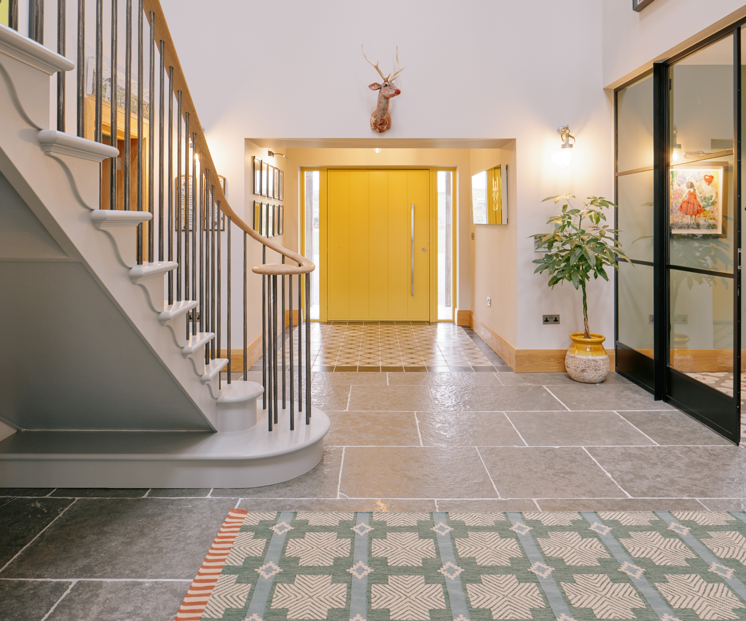Hallway with curved staircase, yellow front door, patterned rugs, stag head and stone flagstone floor.