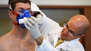 a shirtless man on the left of the photo stands still as an older man wearing a white coat, glasses and gloves adjusts a breathing apparatus on the first man's face