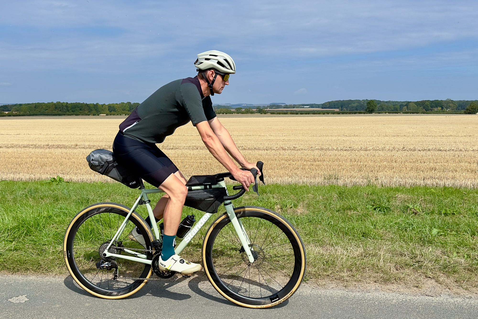 Man riding a green Pfadfinder bike next to an arable field with a green jersey on and bikepacking bags
