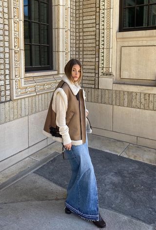 A denim skirt outfit is shown in a photo of a woman on the sidewalk wearing a tan shearling vest over a white long-sleeve sweater with a denim maxi skirt and brown knee-high boots