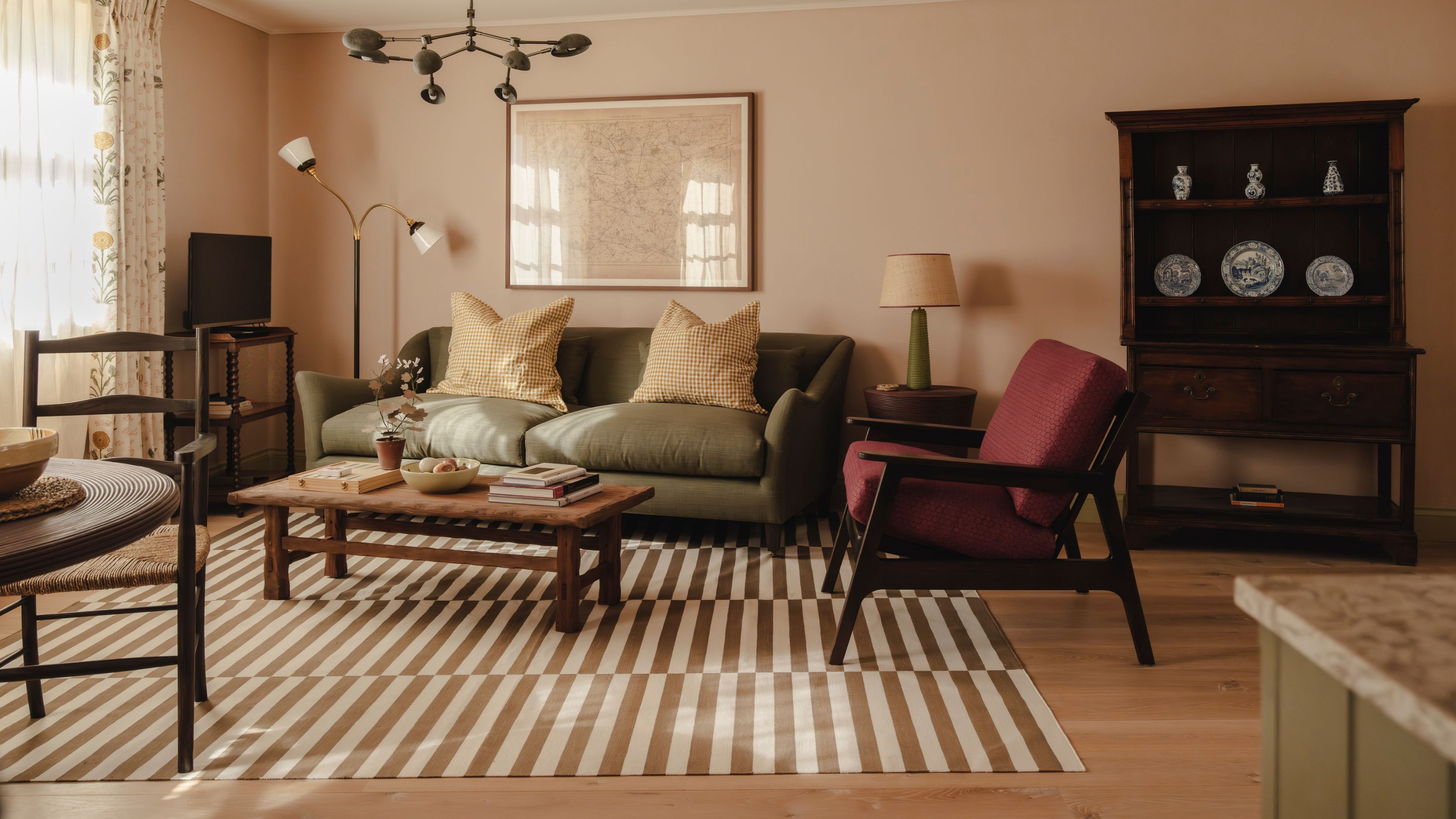 a soft pink living room in the cotswolds with a small olive green couch, burgundy armchair, dark wooden vintage furniture and an ochre striped rug