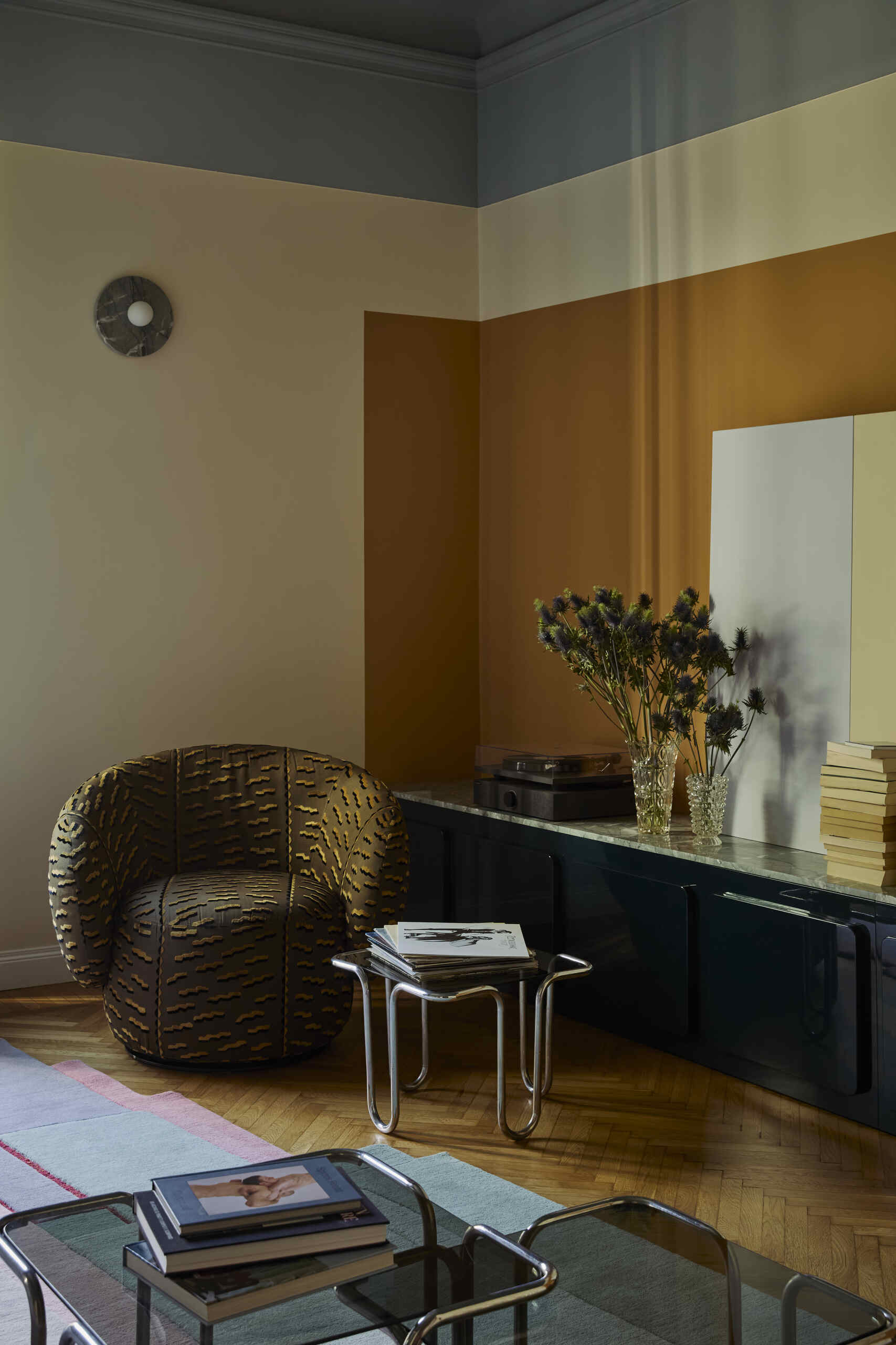 The corner of a luxurious living room with walls painted in a color-blocked orange, white, and blue colorway. There is a tiger-print chair, a record player, and three chrome coffee/side tables.