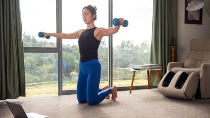 A woman in an apartment exercises on her knees with dumbbells. She is kneeling, with her body erect, and a pair of light dumbbells held out at her sides. Behind her wee see a reclined chair and a large glass window overlooking a green field.