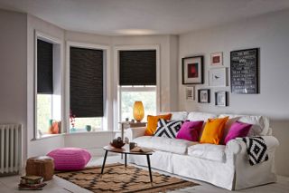 white scheme living room with black out blinds in bay window, white sofa styled with colourful cushions and a gallery wall