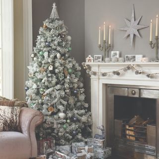 A living room with a fireplace and and a snow-covered artificial Christmas tree in the corner with decorations