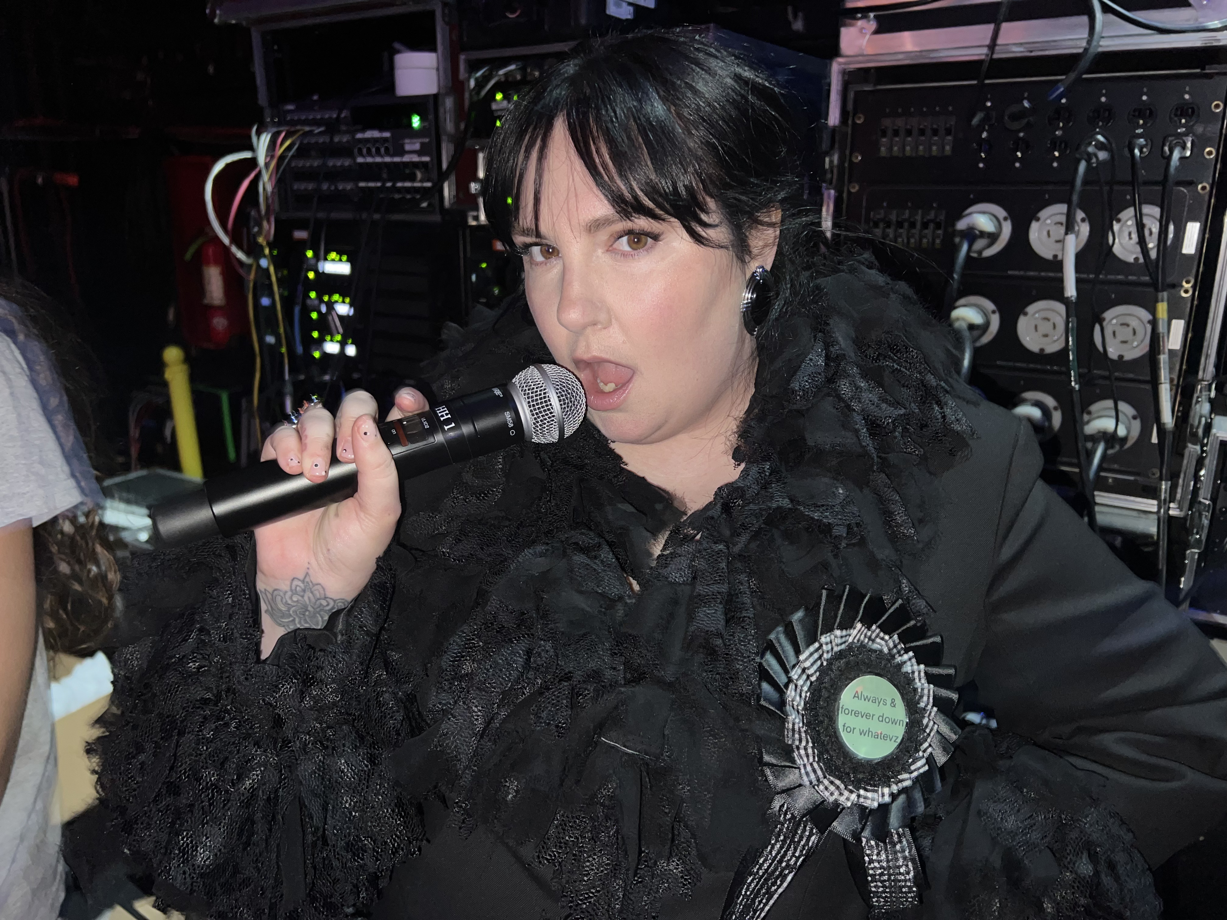 Lena Dunham holding a microphone wearing a black shredded lace blazer and prize ribbon brooch