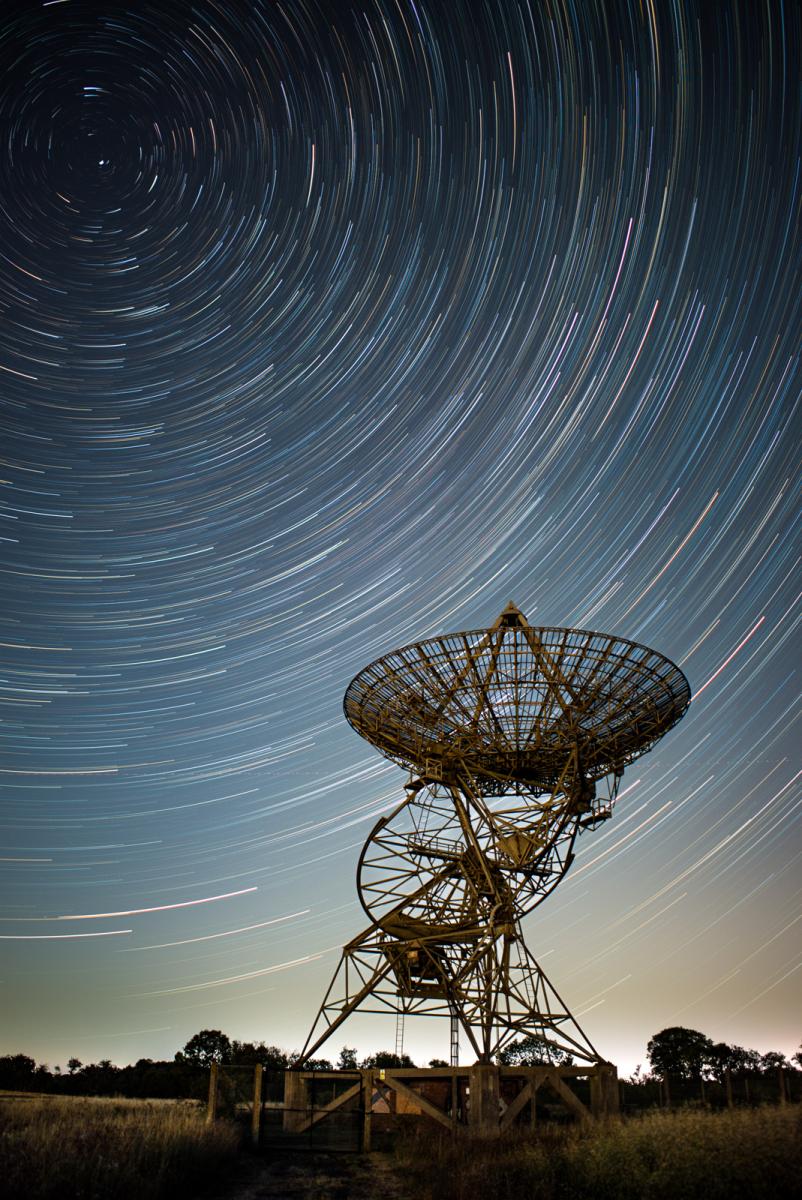 A deactivated radio telescope array sits beneath a circular swirl of stars, exposed to show the motion in the sky as the Earth rotates.