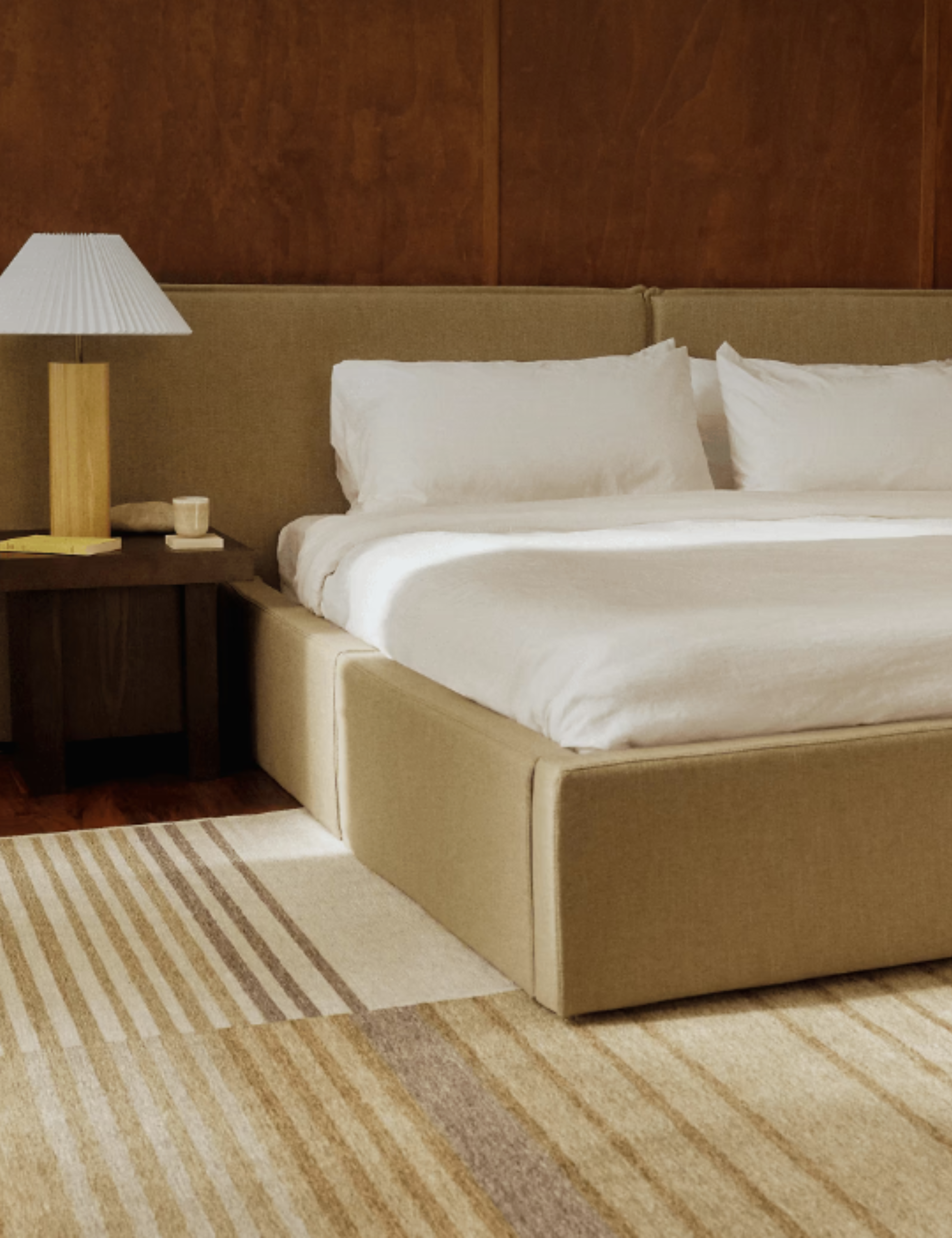 Beige Cozey bed frame with extended headboard featured in a warm wood-paneled bedroom alongside a lamp with a pleated shade and geometric rug