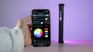 Lume Cube RGB Tube Light Mini in the backgound emitting a purple light, with hand holding phone in the foreground displaying the Lume Control app