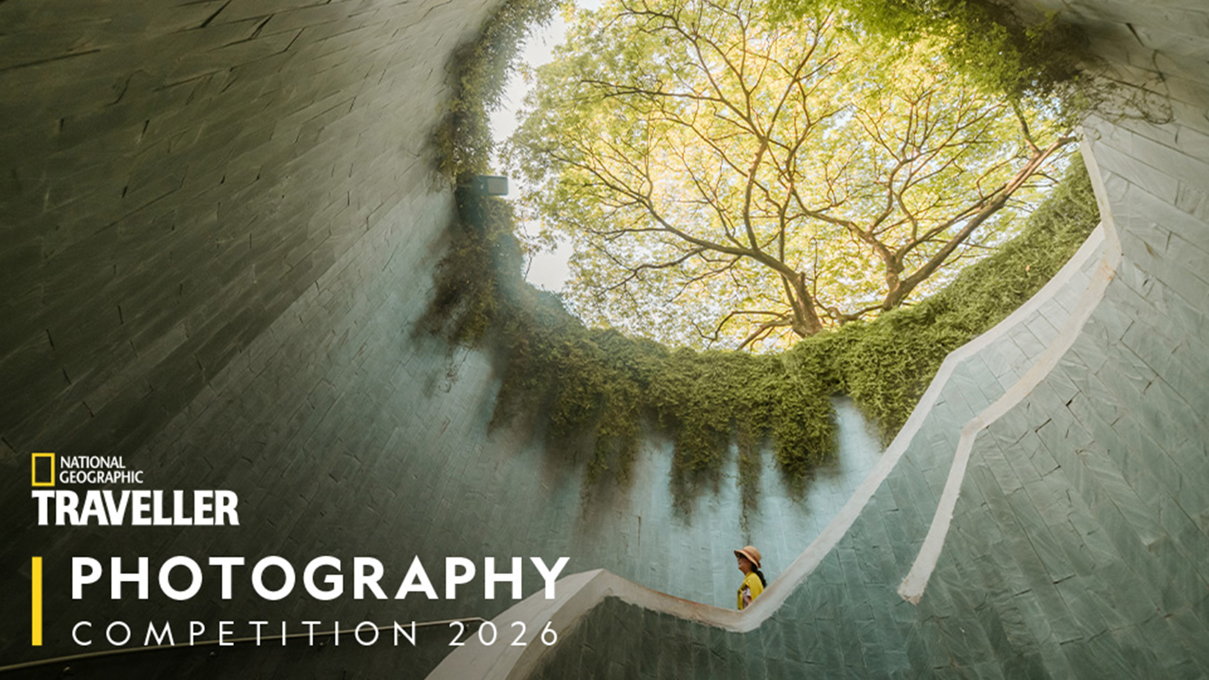 A person stands on a white spiral staircase, surrounded by tall green walls and a view of trees above, creating a serene atmosphere