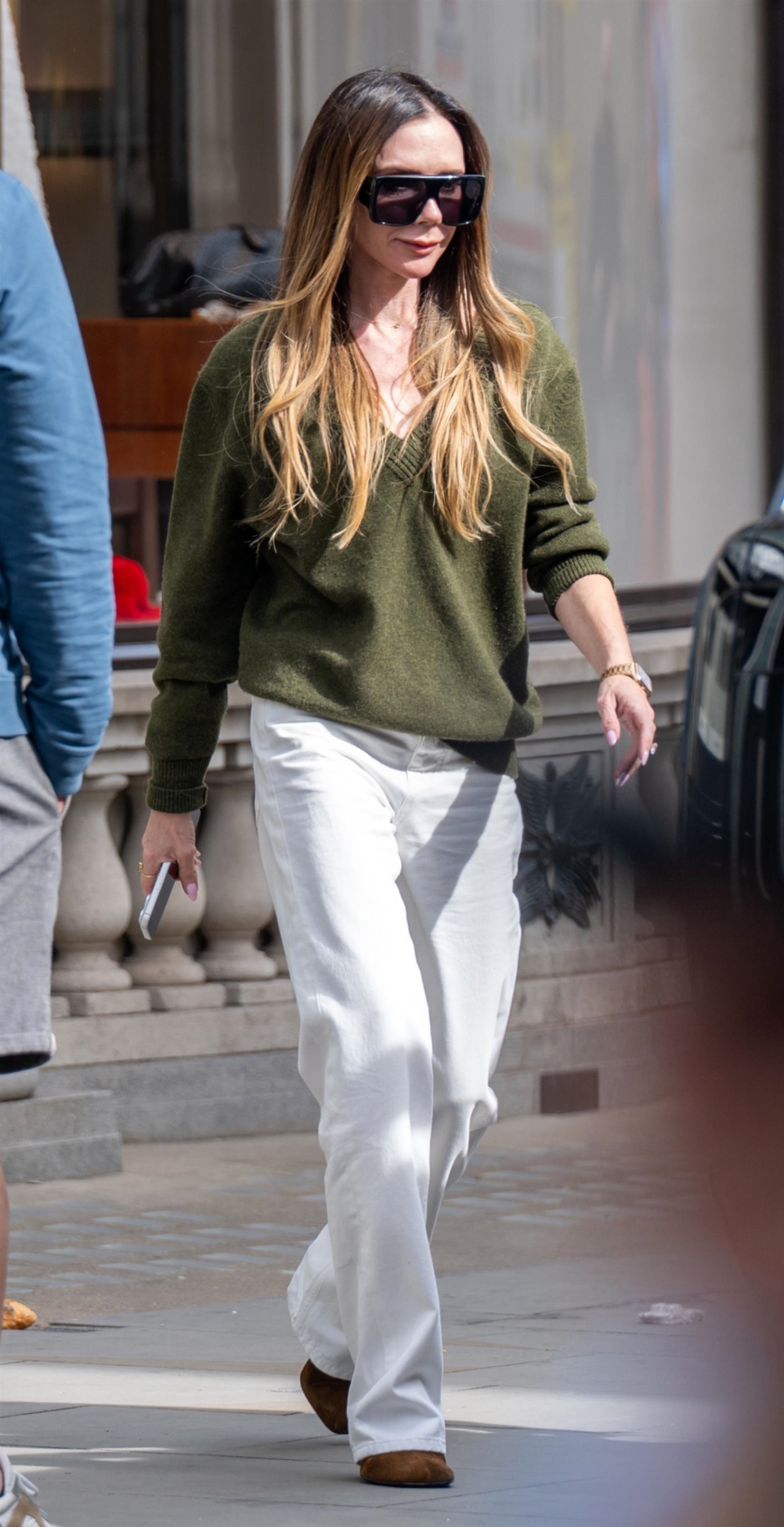 Victoria Beckham wears white jeans with an olive v-neck jumper and brown suede shoes.