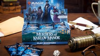 A Booster Display box for Murders at Karlov Manor sitting on a wooden table alongside bronze curiosities, dice, and other props