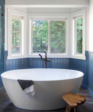A bathroom with four windows, cream white walls, dark blue wooden wall panelling, and a white curved bath tub with a dark brown wooden stool next to it