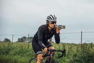 A cyclist is drinking from a bottle while riding a bike, in the background is a field and grey sky.