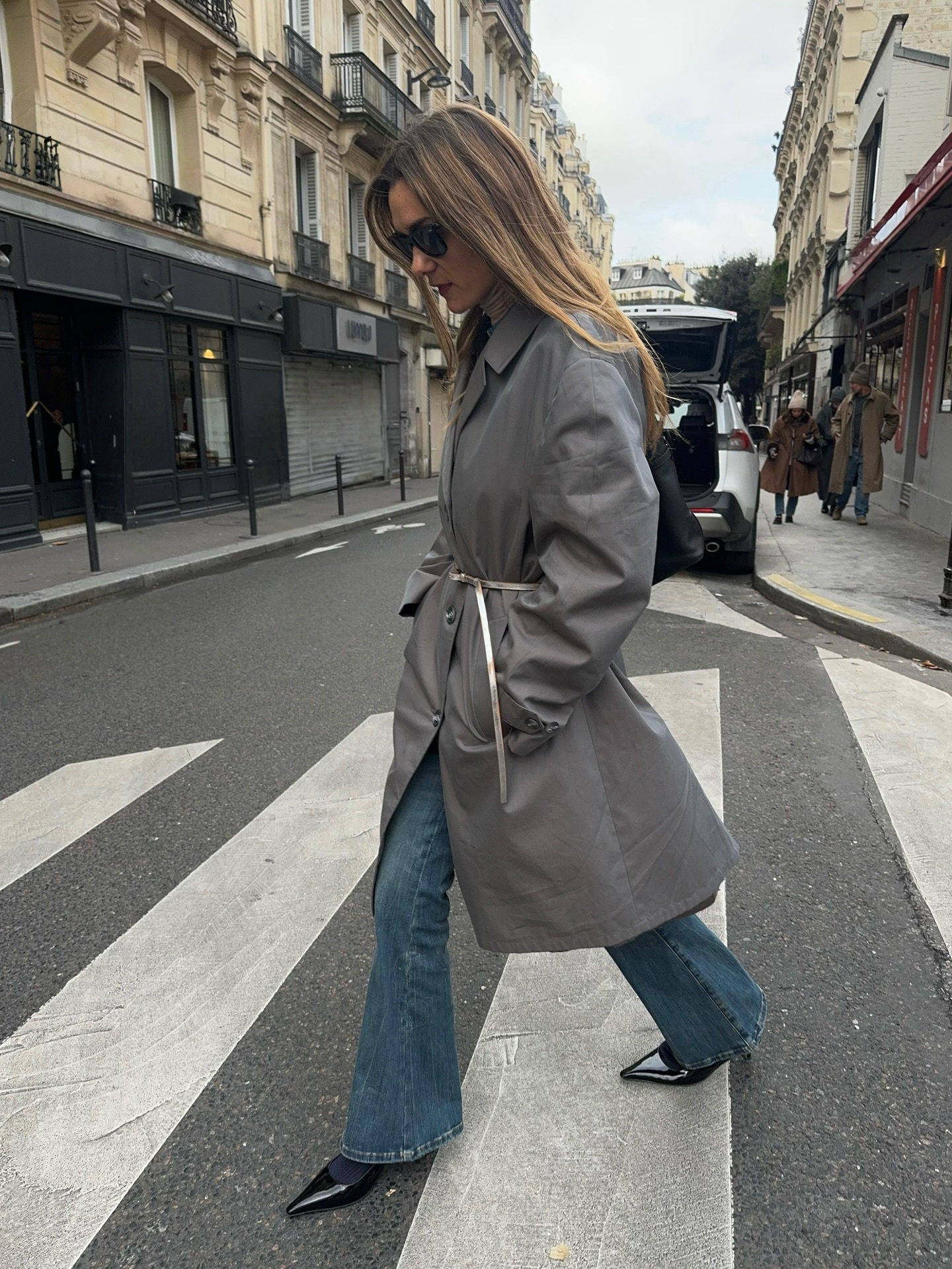 An image of an influencer in Paris wearing a grey trench coat with a silver belt with flare jeans and black pointed-toe patent leather heels