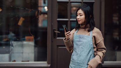 A small business owner stands outside her business and checks her investments on her smartphone. 