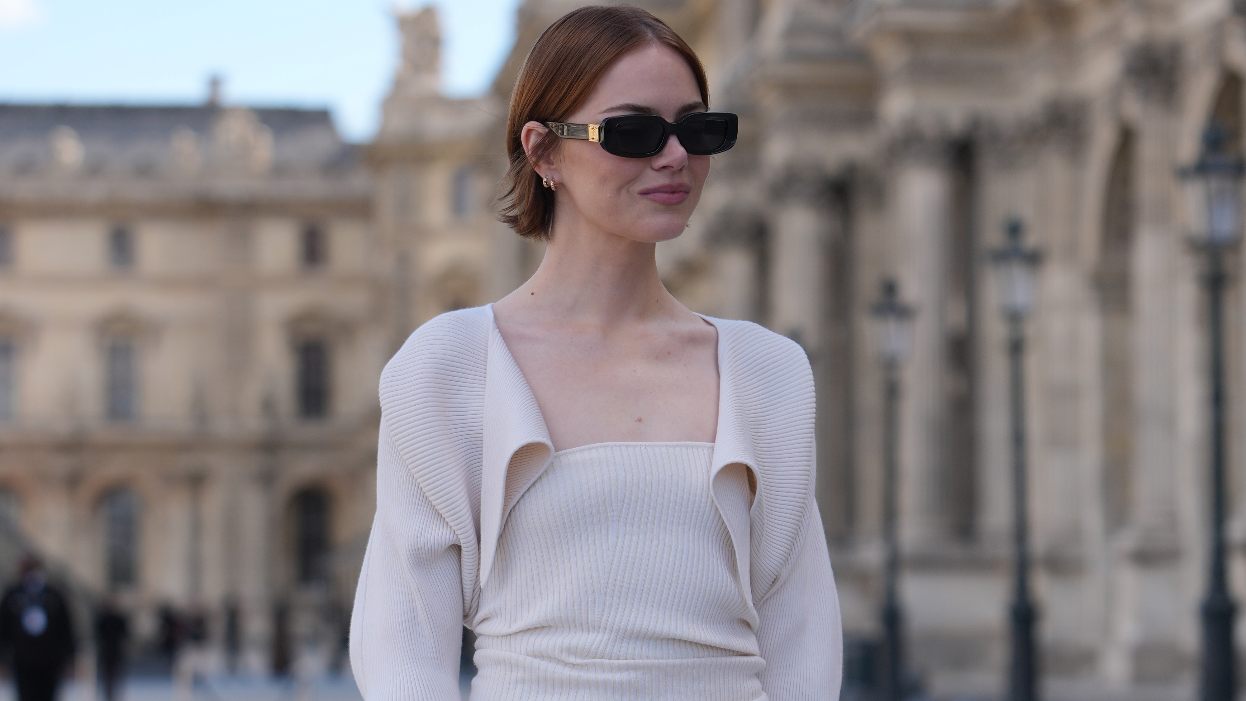 PARIS, FRANCE - SEPTEMBER 30: Emma Stone wears auburn chin-length straight hair with a center part, black rectangular sunglasses with gold metal temple detailing, gold hoop earrings and a gold ring, a black leather satchel bag with top handles and silver hardware carried handheld, a cream ribbed knit strapless midi dress with a ruched bodice and fitted pencil skirt layered with a matching cream ribbed knit bolero-style cropped long-sleeve cardigan with an open draped front, black leather pointed-toe pumps shoes with stiletto heels, outside Louis Vuitton, during Paris Fashion Week Womenswear Spring/Summer 2026, on September 30, 2025 in Paris, France 