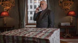 Brian Cox takes a phone call while standing in front of a huge money pile in 007: Road to a Million.