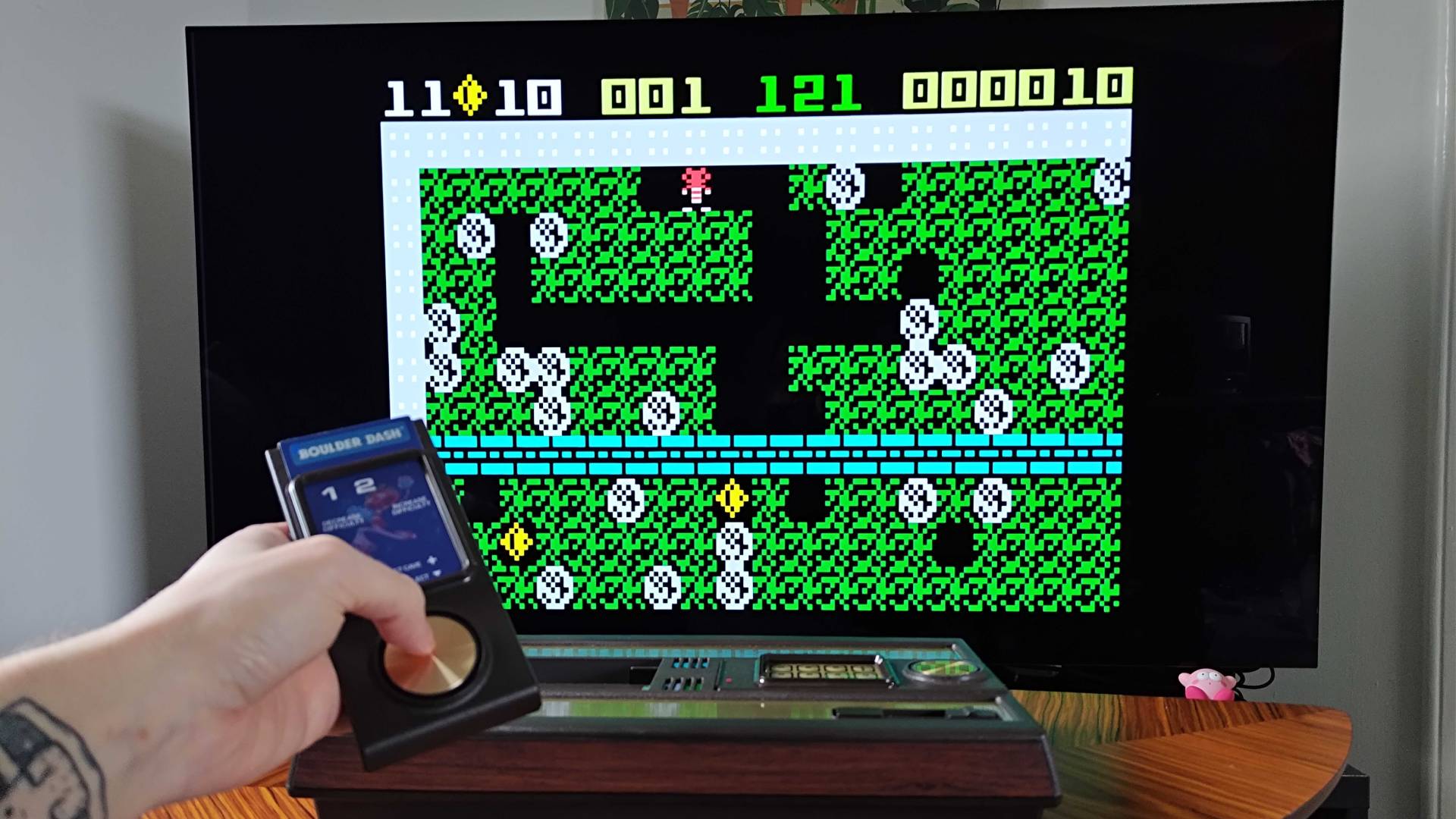 Hand holding Intellivision Sprint controller with Boulder Dash gameplay on TV in backdrop.