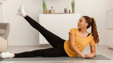 Woman performs side leg lift exercise at home. She is lying on her side on a black exercise mat, propping her torso up on a forearm, with her top leg lifted and extended. She is wearing white trainers, black athletic leggings and a yellow short-sleeved top.