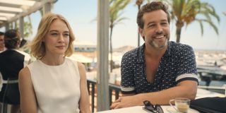 Elen Rhys and Julian Looman as Miranda Blake and Max Winter in The Mallorca Files S3. They are both sitting on the same side of a table in the open-air section of a cafe. Max is leaning on the table and smiling.