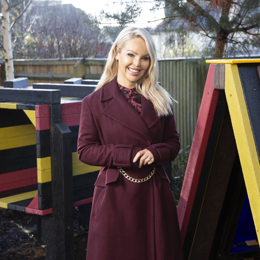 Katie Piper poses for a photo in a garden while wearing a a purple coat