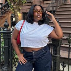 Editor Aniyah Morinia wears white t-shirt, jeans, red handbag and eyeglasses while standing in front of New York City home.