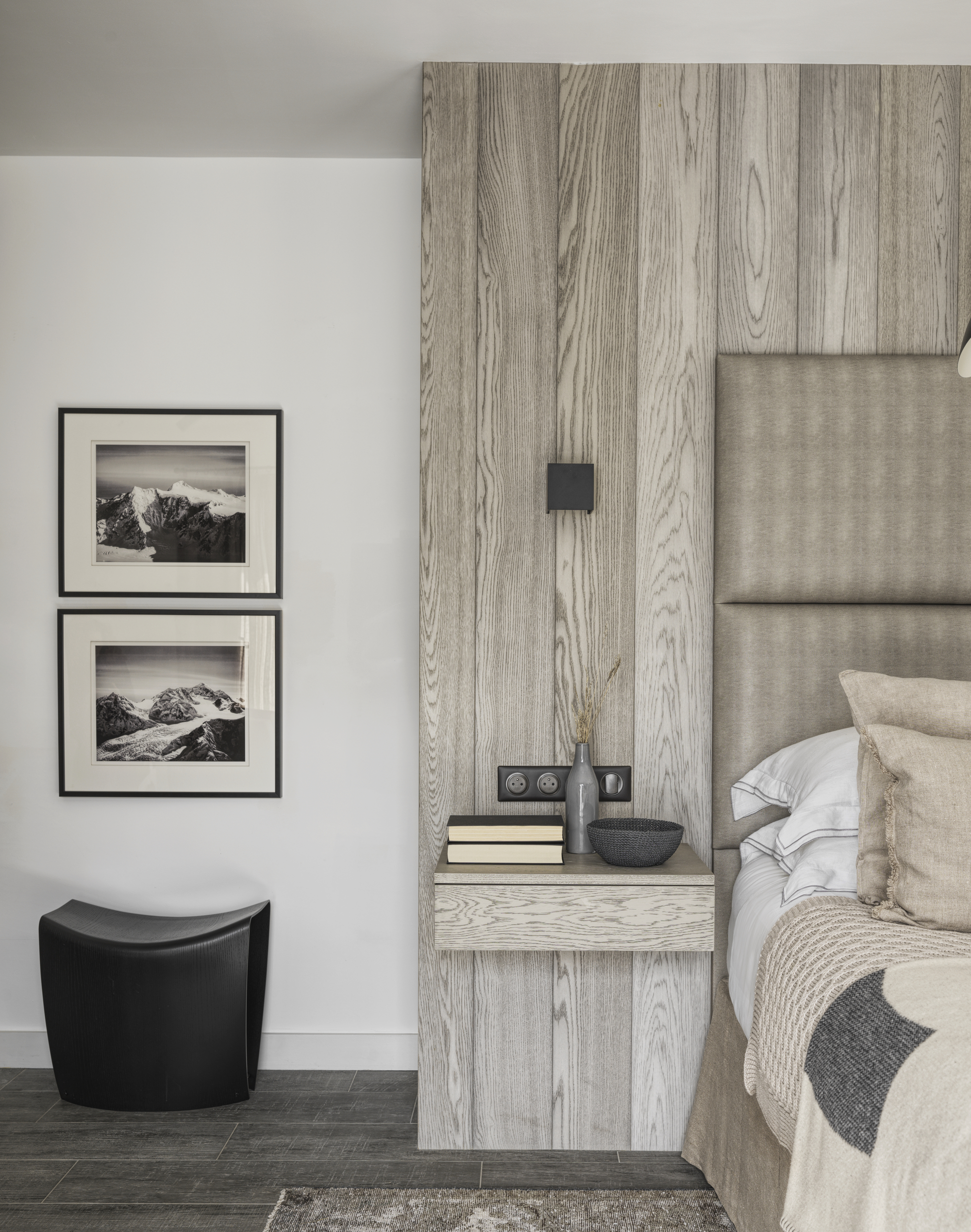 neutral bedroom with timber paneling around bed with built-in floating side tables styled with books and bowl, neutral and white bed linen, beige upholstered bedhead, dark gray flooring, black ottoman underneath two black and white framed pictures on white wall