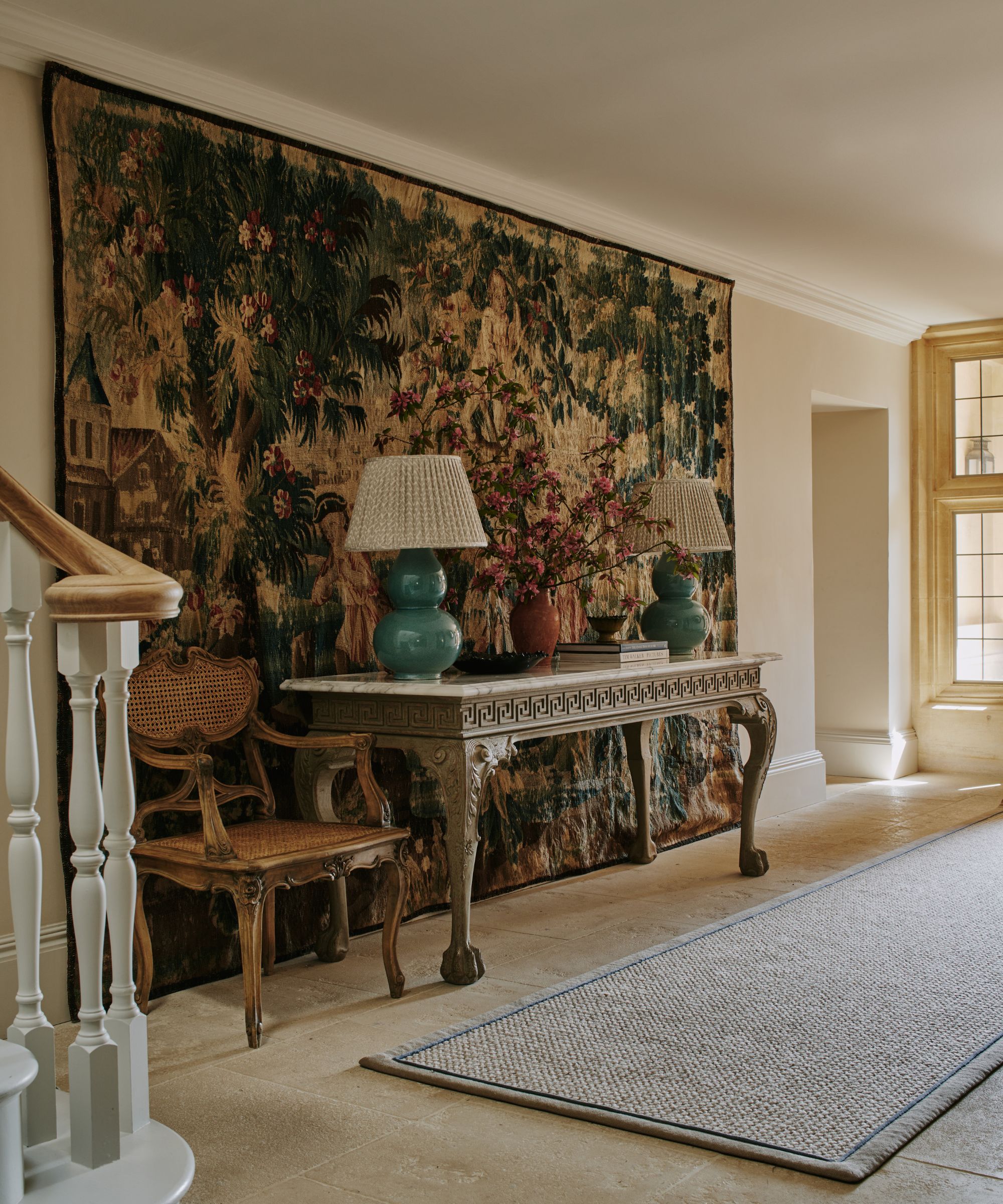 Entryway with antique iron and marble console, rococo wooden chair, large antique tapestry hanging on the wall behind and two teal lamps on the table with a long jute runner on the floor
