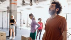 Man stands with slightly apprehensive look on his face. He is in a gym and three women stand in the background next to wooden plyo boxes