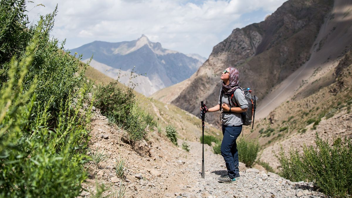 A woman wearing Montane Women&#039;s Terra Stretch Lite Trousers and holding trekking poles stands on a stony mountainside, looking up.