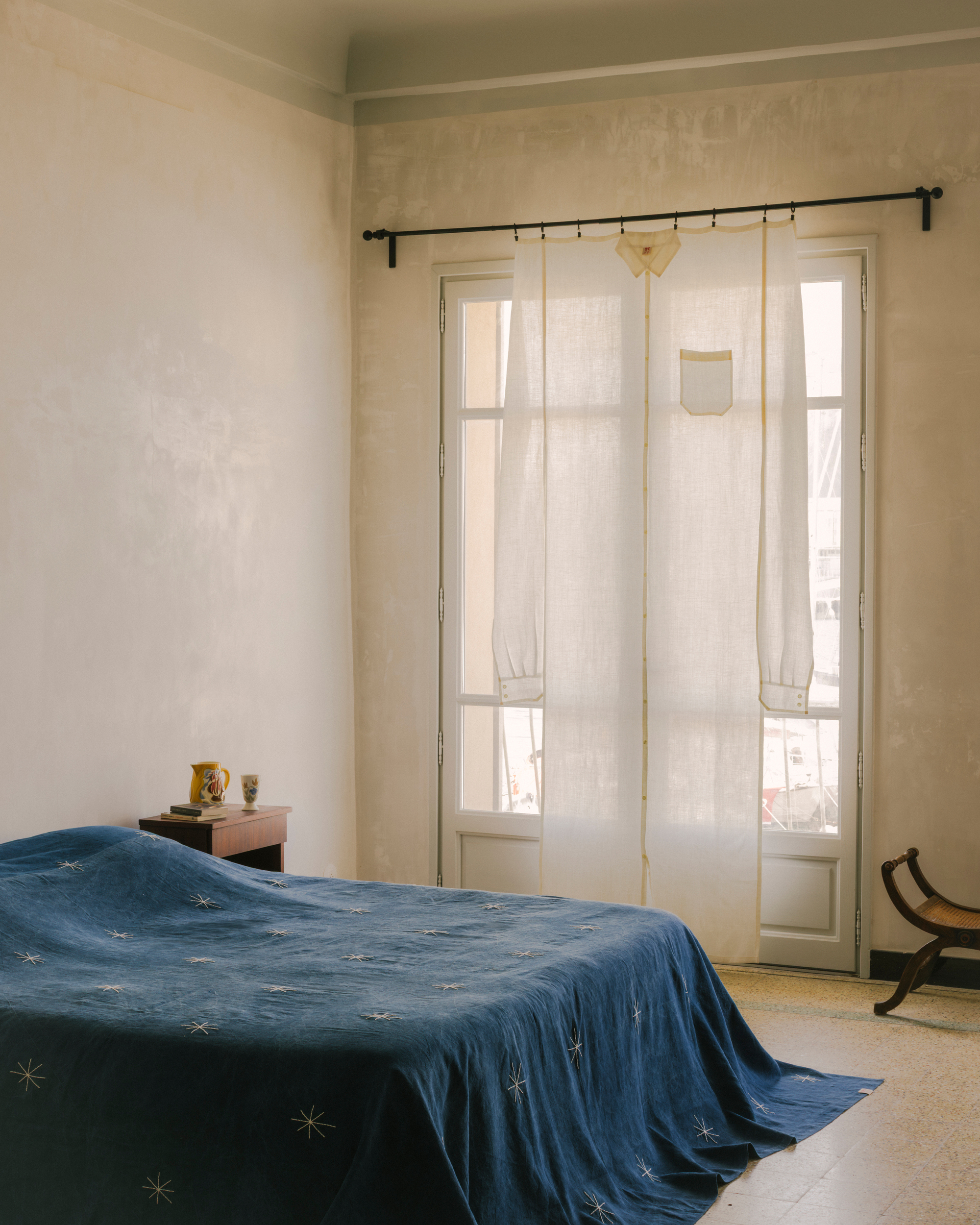 A bright, lived-in bedroom with a minimalist bed that has a navy blue bed cover with embroidered stars on it. There are French doors with a noren-style curtain hanging in front of them that's shaped like a button-down shirt.