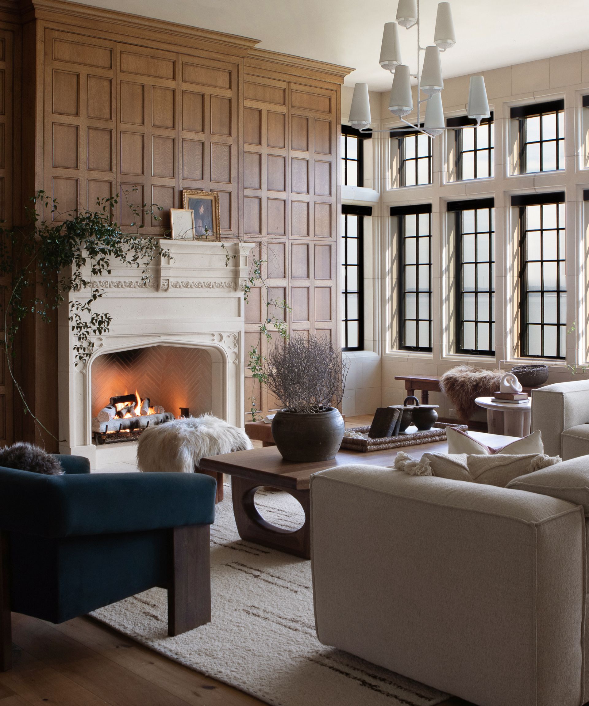 winter decor ideas, cozy living room with panelled walls, open fire lit, armchairs, wooden coffee table, cream couch, cream rug, faux fur/sheepskin, white retro pendant light