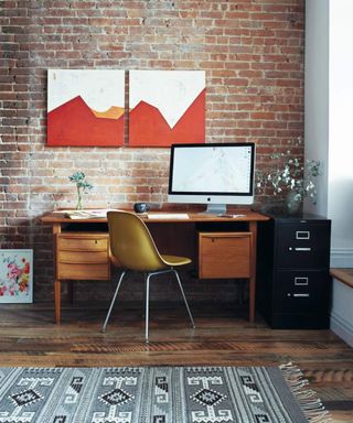 A home office with dark wood desk and yellow chair with macbook in front of a brick wall