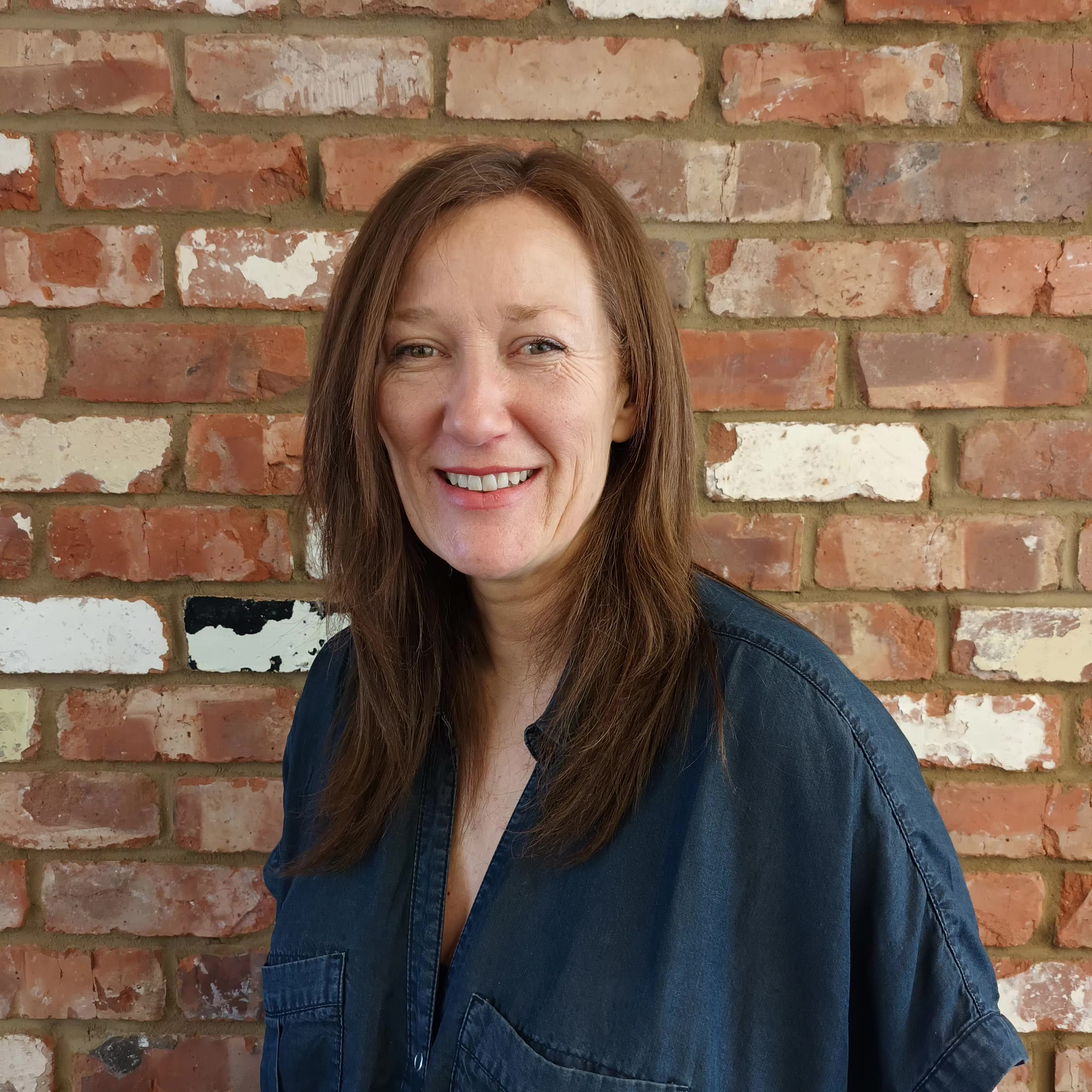 A woman with brown hair and a dark blue top against a redbrick wall