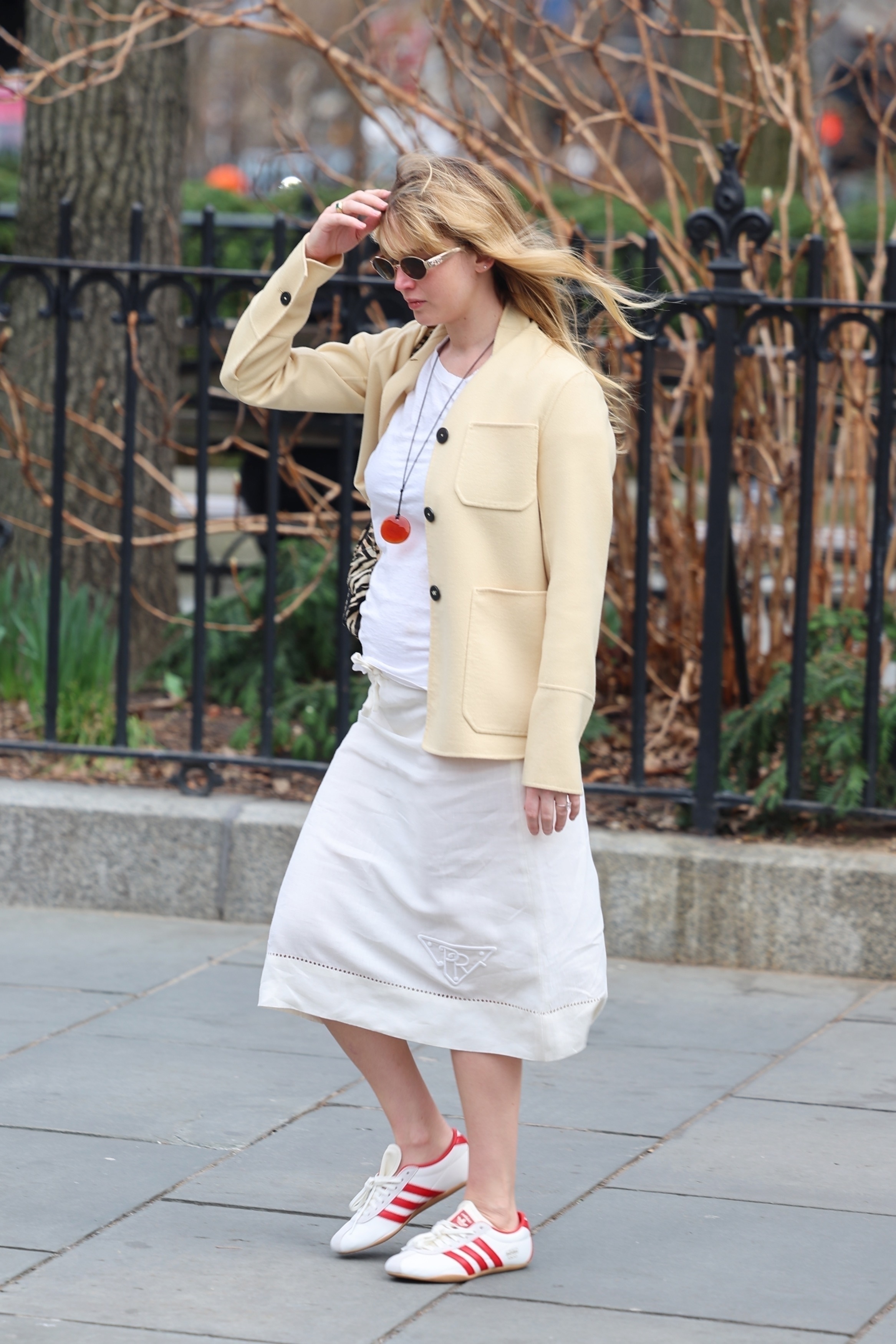 Jennifer Lawrence pictured in a pair of Adidas Tokyo trainers, a cream Prada midi skirt, a white top and a cashmere butter yellow jacket. She accessorises the look with a red rope necklace and Celine Triomphe sunglasses in a recent image taken of her.