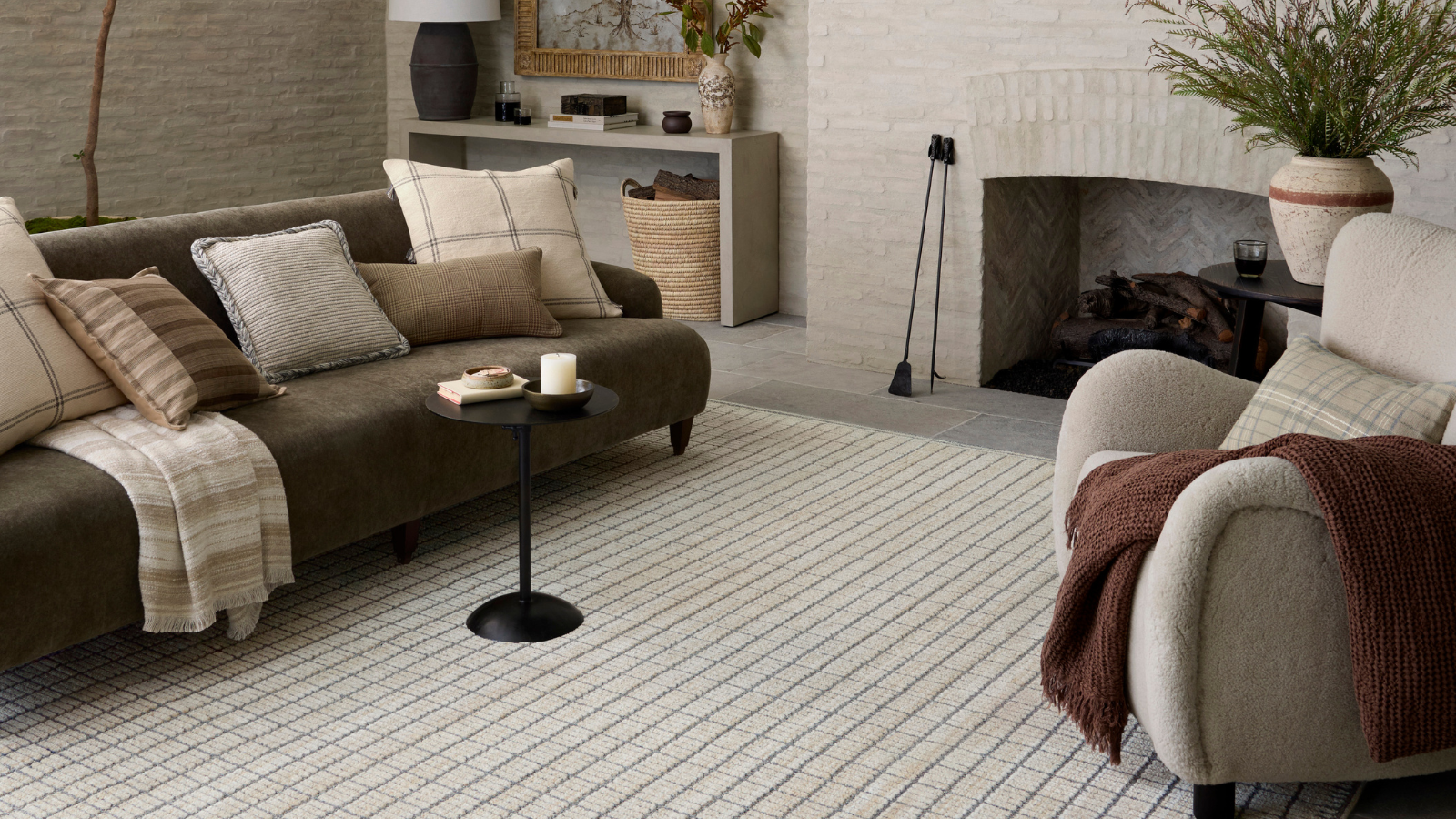 a neutral colored living room with a brown couch covered with pillows, a small black drink table, an accent chair with a brown blanket, a checker printed rug, and a large white-washed brick fireplace in the background