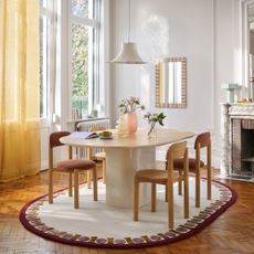 An open-plan dining room with an oval dining table on a cream oval rug with a contrasting border and a yellow curtain on high windows 